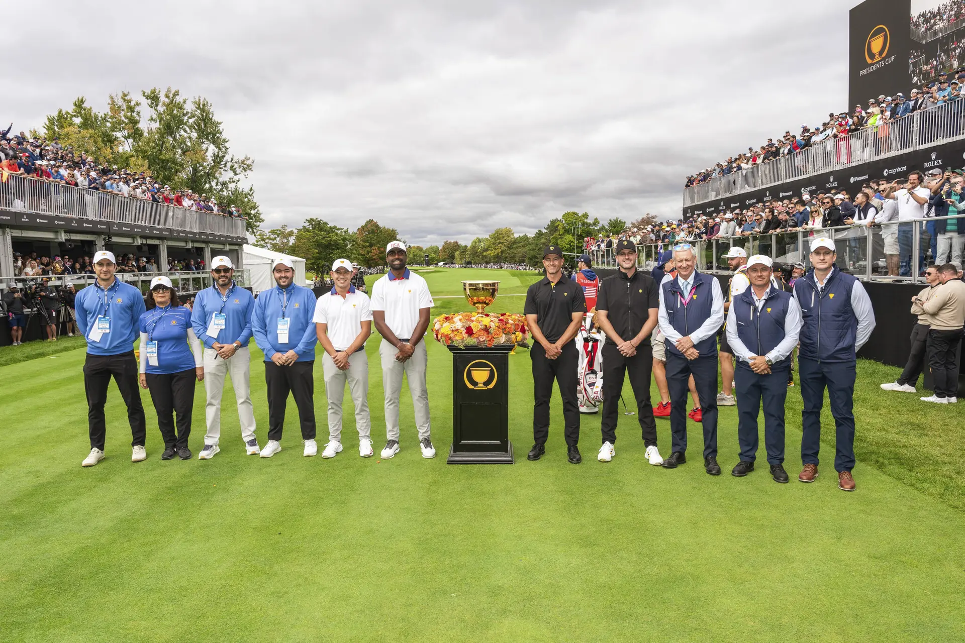Golf players and officials standing on a golf course green around a trophy with spectators in stands behind them.