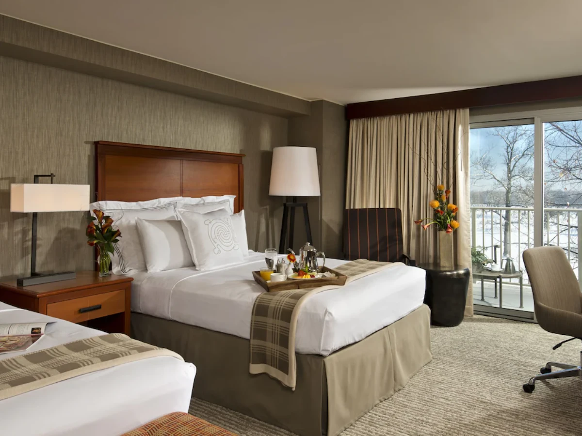 Hotel room with two beds, a wooden nightstand with a lamp and flowers, a tray with breakfast on one bed, an armchair, floor lamp, and sliding glass doors leading to a balcony overlooking snowy trees.