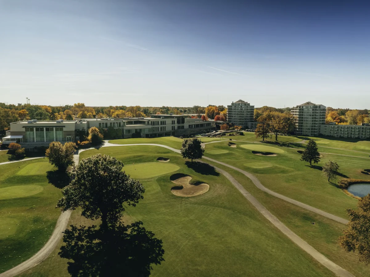 Aerial view of a golf course with bunkers, trees, pathways, and multiple buildings in the background under a clear blue sky.
