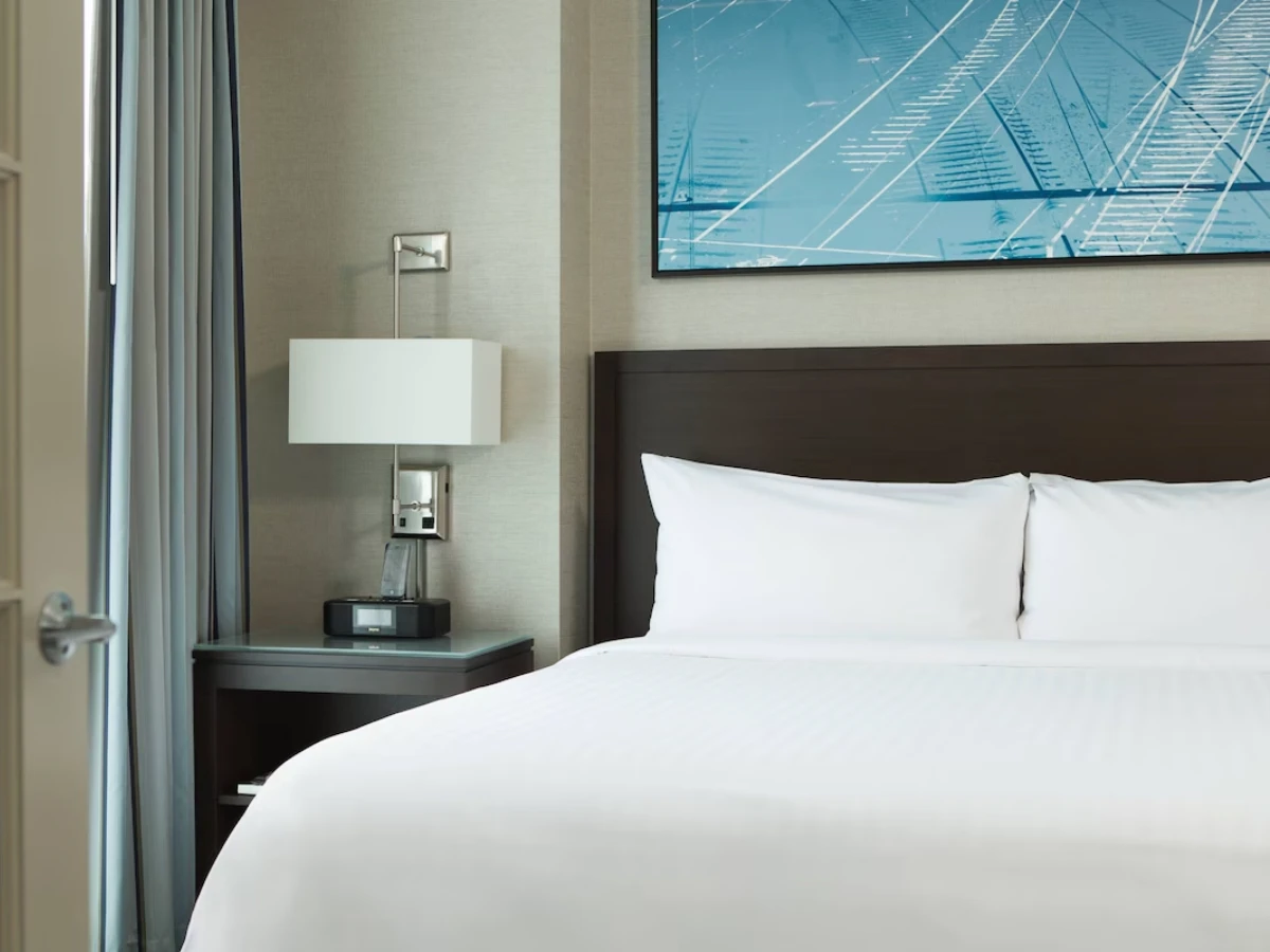 Hotel room with a neatly made bed with white linens, a dark wood headboard, a nightstand with a lamp and alarm clock, and a blue abstract painting above the bed.