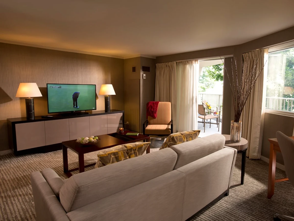 Living room with beige sofa, two patterned pillows, armchair, coffee table, TV on a console, two lamps, and sliding glass doors leading to an outdoor balcony with chairs and greenery.