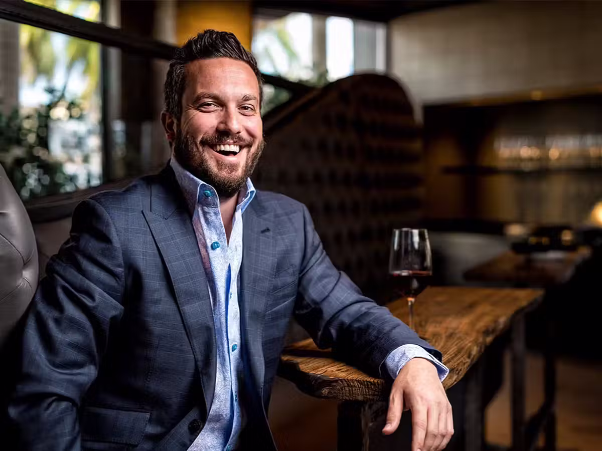 Smiling man in a blue suit jacket sitting at a wooden bar table with a glass of red wine.