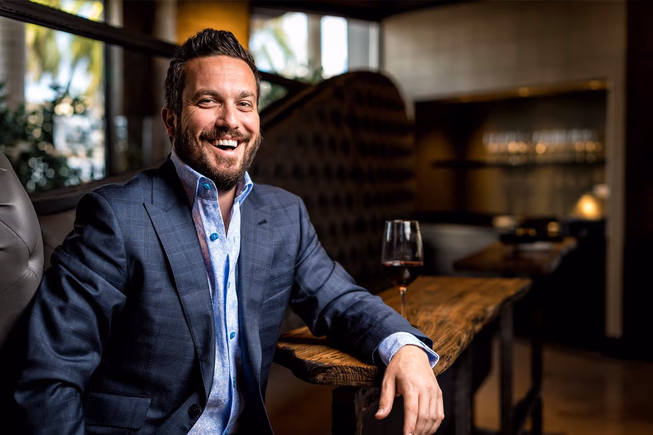Smiling man in a blue suit jacket sitting at a wooden bar table with a glass of red wine.
