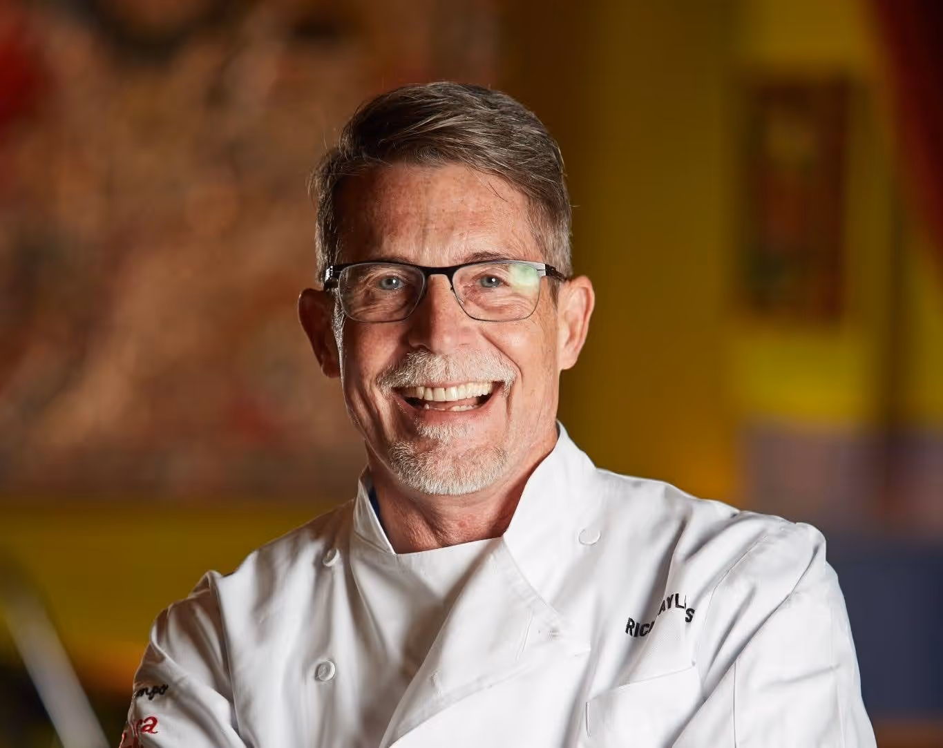 Smiling middle-aged man wearing glasses and a white chef's coat with crossed arms.