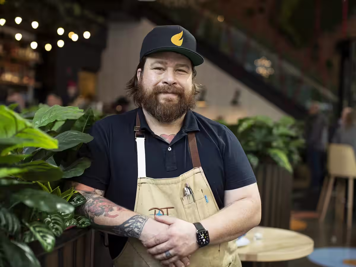 Bearded man wearing a navy cap and apron standing with arms crossed in a cozy indoor space with plants and warm lighting.