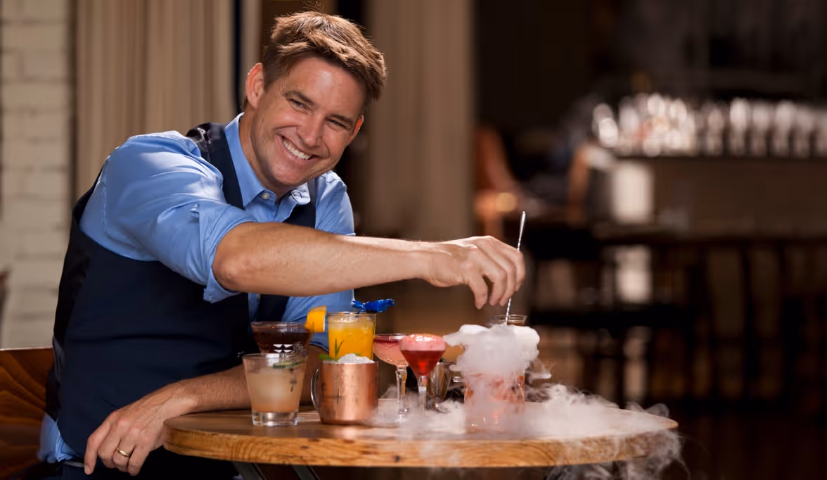 Man in blue shirt and black vest stirring a smoking cocktail at a wooden table with various colorful drinks.