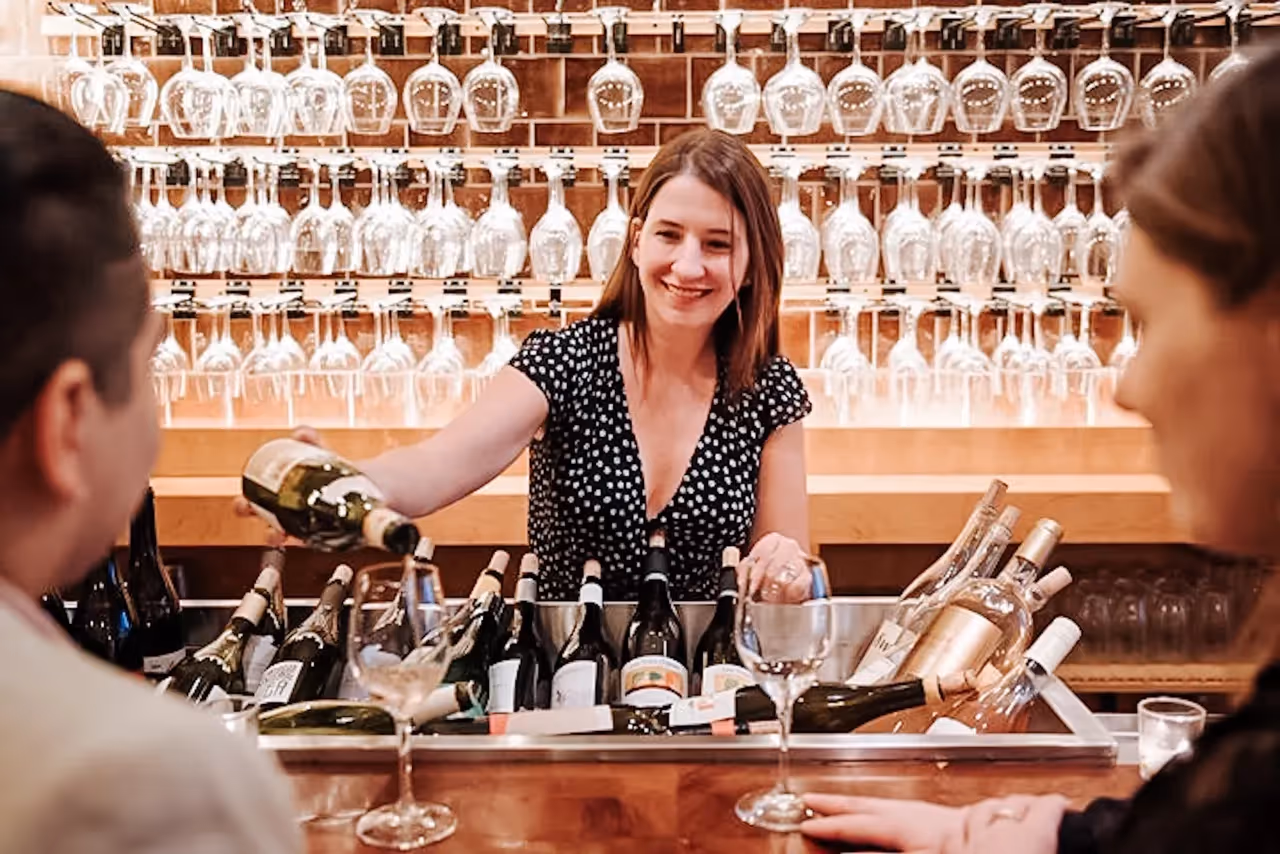 Smiling bartender pouring white wine into a glass with wine bottles and hanging glasses in the background.