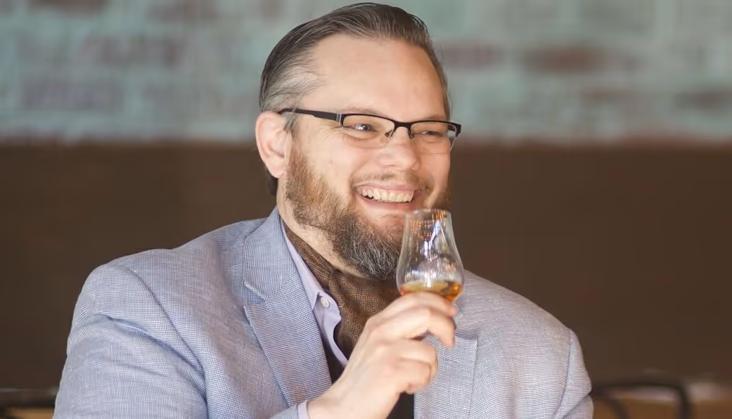 Smiling man wearing glasses and a light gray blazer holding a glass of amber liquid, possibly whiskey, in a dimly lit setting.