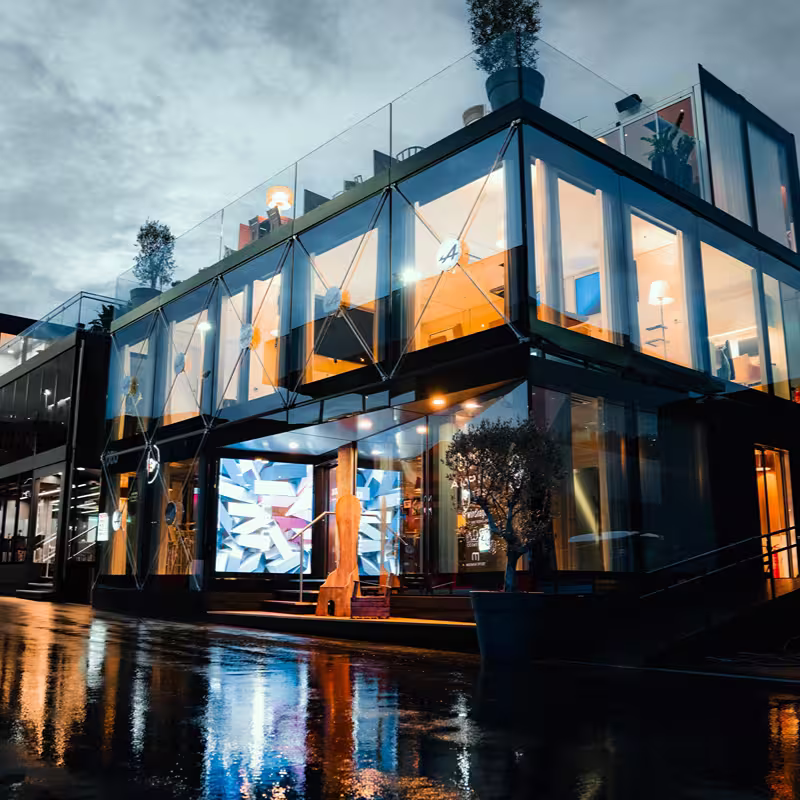 Modern glass building with illuminated interior and rooftop plants reflecting on wet pavement at dusk.