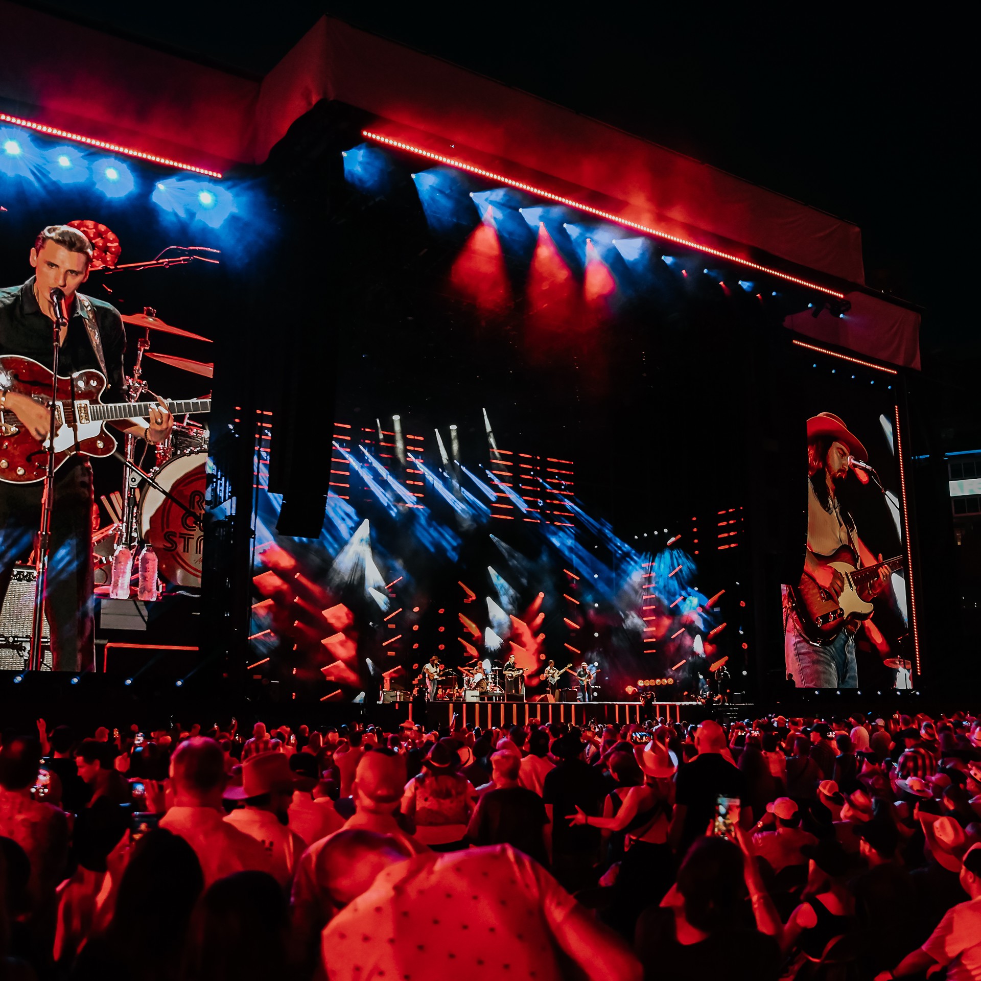 Nighttime outdoor concert with a large crowd watching a band perform on a stage with colorful red and blue lighting and big screens showing close-ups of the musicians.