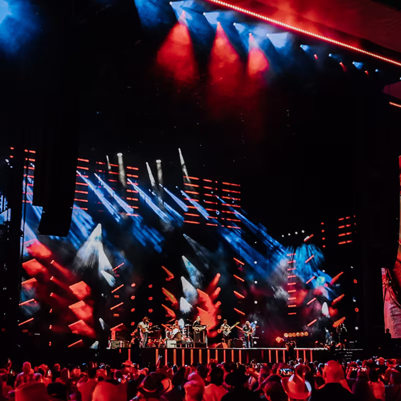Nighttime outdoor concert with a large crowd watching a band perform on a stage with colorful red and blue lighting and big screens showing close-ups of the musicians.