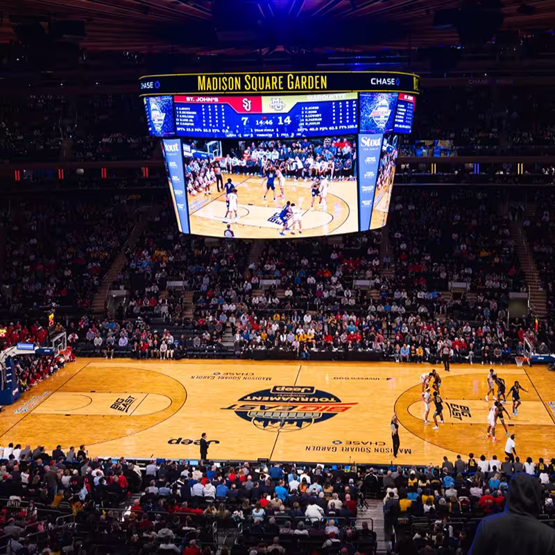 Indoor basketball game at Madison Square Garden with players on the court and a large scoreboard showing St. John's vs Marquette and a crowd watching.