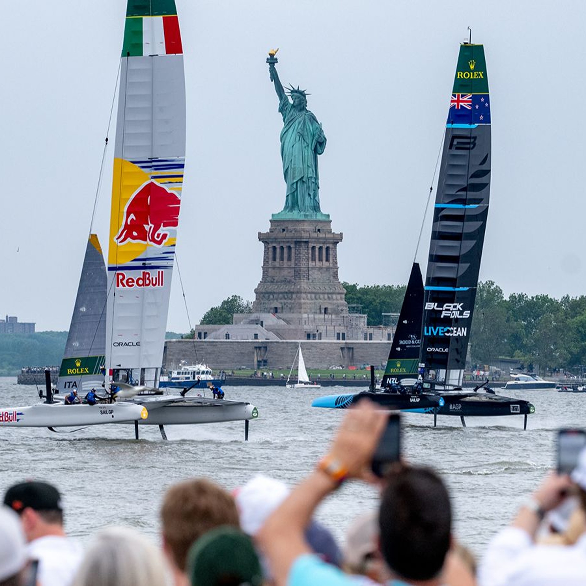 Two sailboats with Red Bull and Black Foils branding racing on the water near the Statue of Liberty while spectators watch and take photos.