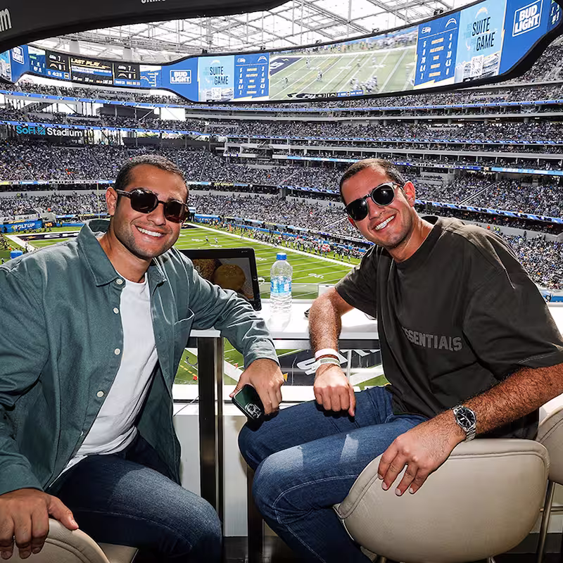 Two men wearing sunglasses smiling and sitting in a stadium suite overlooking a football game at SoFi Stadium.