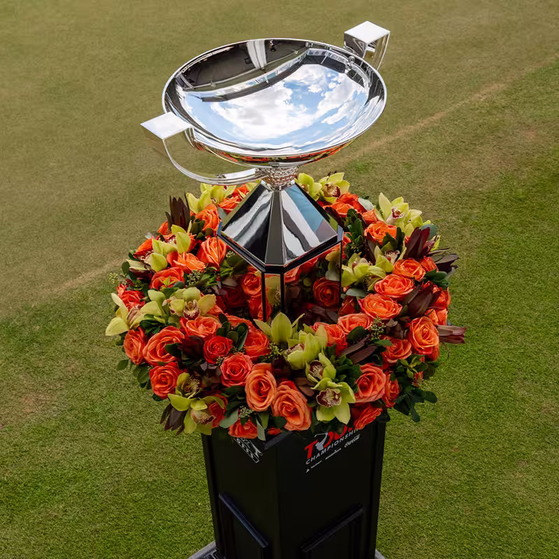 Shiny silver trophy cup surrounded by a wreath of orange roses and green orchids on a grass field.