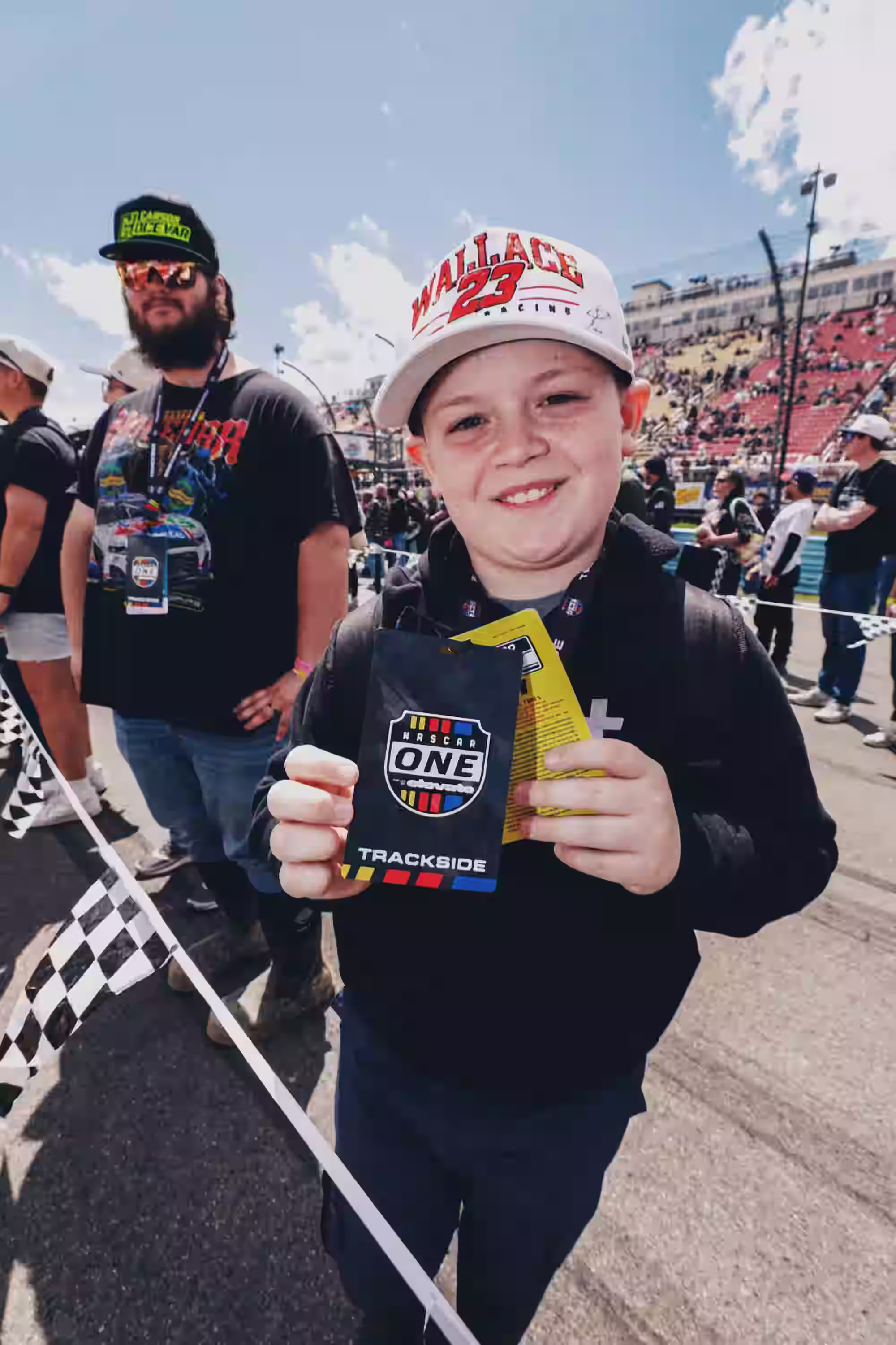 เด็กชายที่ยิ้มแย้มและสวมหมวก Wallace 23 Racing กำลังถือบัตรผ่าน NASCAR One บริเวณข้างสนามในการแข่งขัน