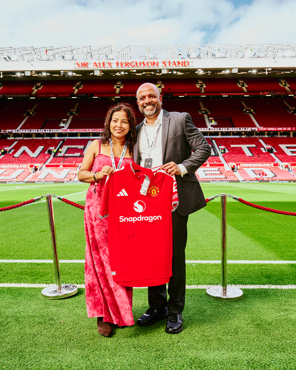 Un homme en costume et une femme en robe rose se tiennent sur un terrain de football, tenant un maillot rouge de Manchester United orné du logo Snapdragon, avec la tribune Sir Alex Ferguson et les sièges rouges du stade en arrière-plan.