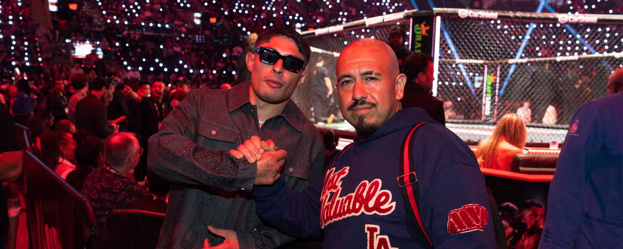 Two men posing for a photo in front of a lit MMA octagon cage with audience in the background, one wearing sunglasses and a dark jacket, the other in a blue hoodie with red lettering.