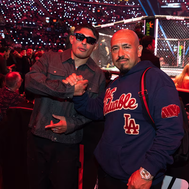 Two men posing together near a UFC fight cage with a crowd in the background, one wearing sunglasses and a dark jacket, the other in a blue hoodie with 'Not Humble' and 'LA' logos.