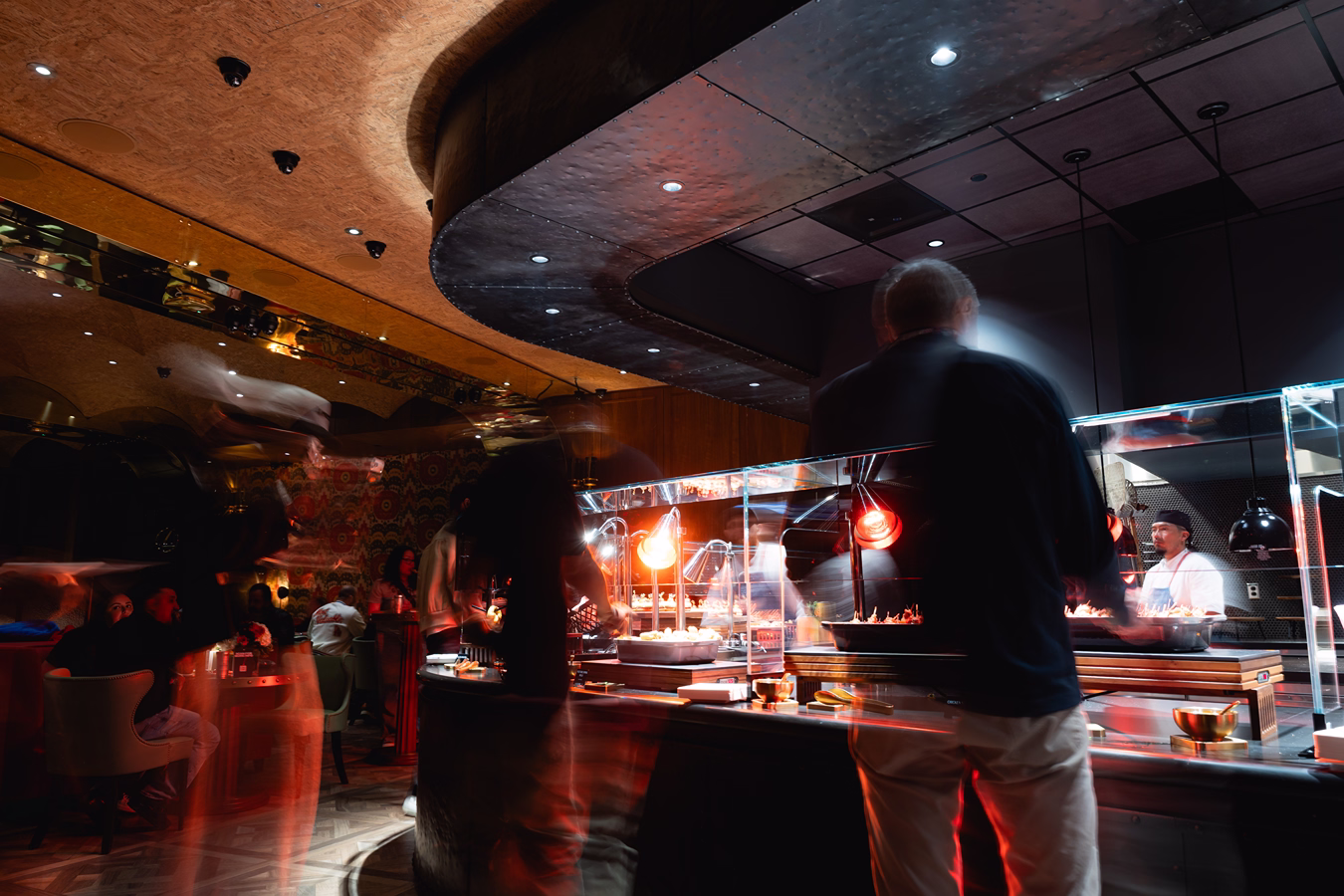 Dimly lit restaurant interior with blurred figures of people moving near a buffet counter featuring heated food lamps and staff behind a glass partition.