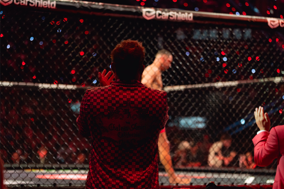 View through the cage of a mixed martial arts event with a shirtless fighter walking inside and two people in the foreground clapping and raising hands.