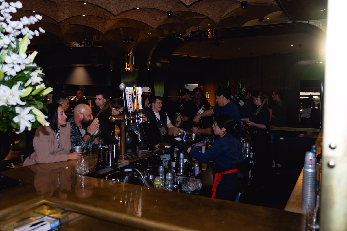 People sitting and standing around a dimly lit bar counter where bartenders are serving drinks, with a bouquet of white flowers visible on the left side.