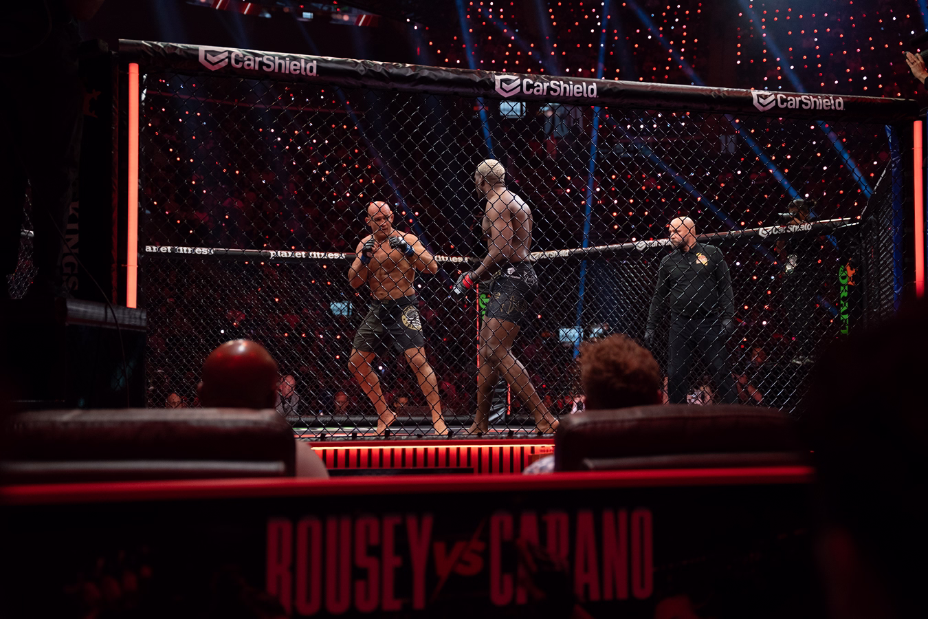 Two mixed martial artists face off inside an octagonal cage during a UFC fight, with a referee standing nearby and an audience visible in the background.