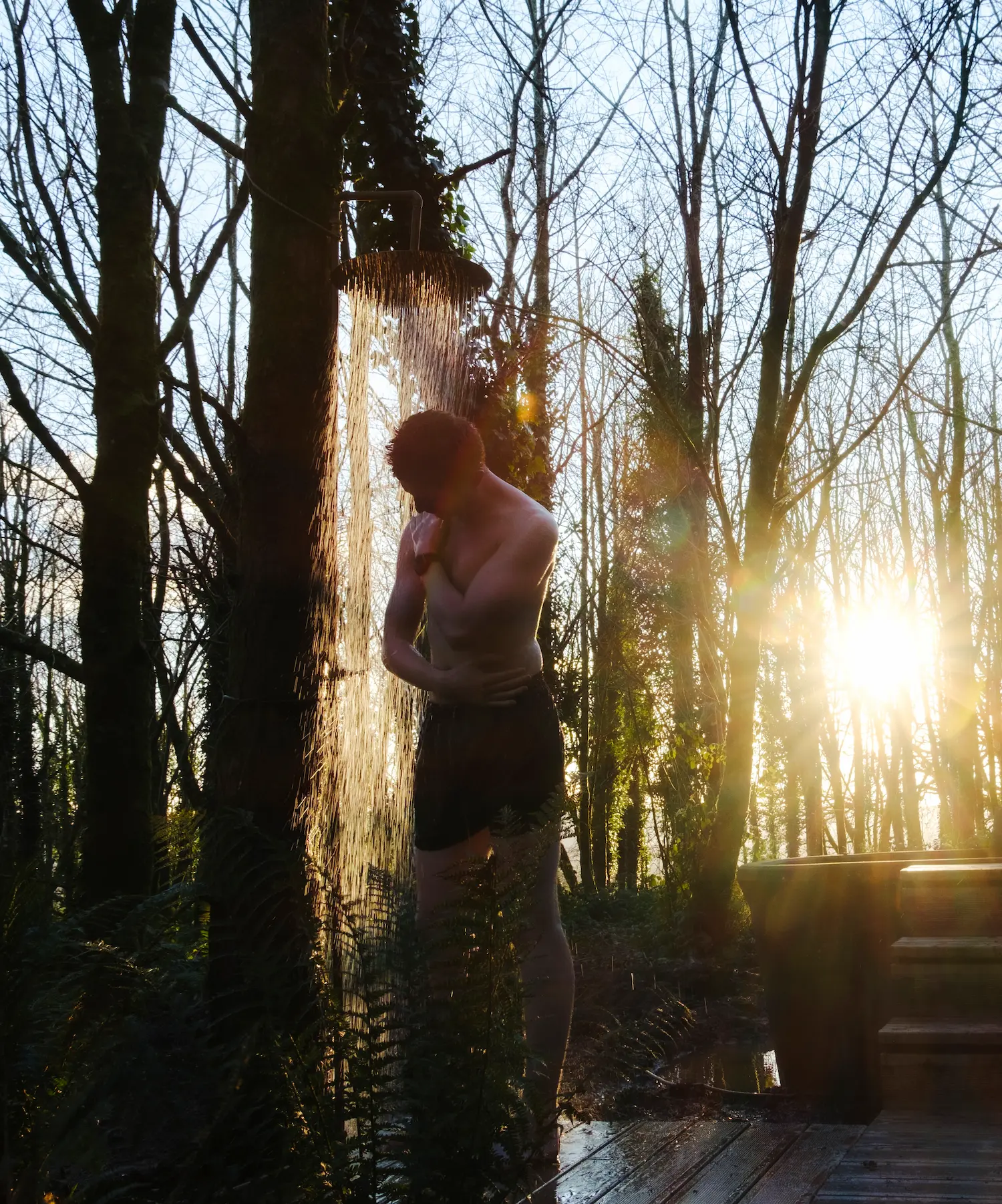 Person standing under an outdoor shower at The Sauna In The Woods in a forest with sunlight streaming through bare trees at sunrise or sunset.
