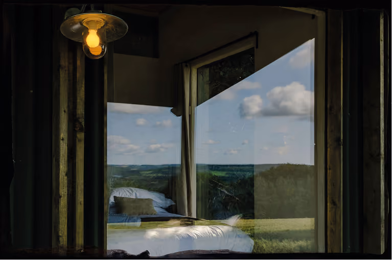 Reflection of bed and landscape outside seen through a window with glowing pendant light above.