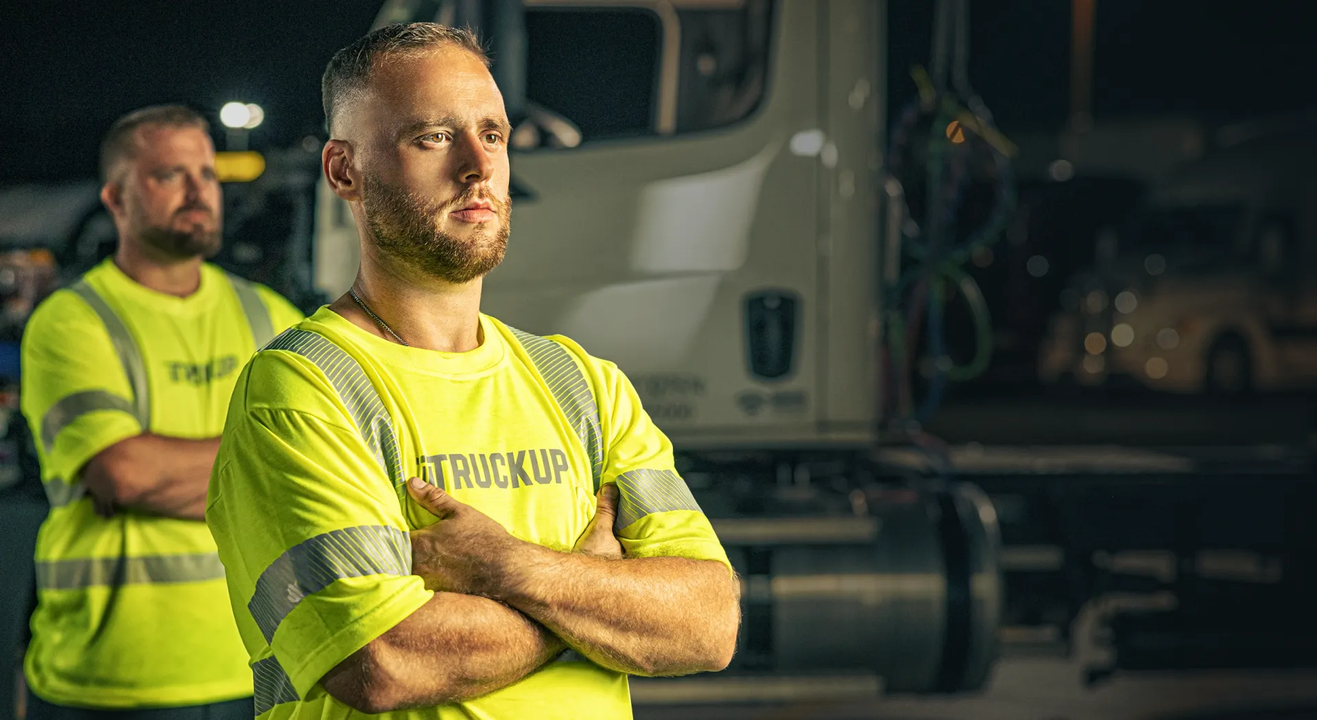 Two men wearing bright yellow safety shirts with reflective stripes standing with arms crossed in front of a truck at night.
