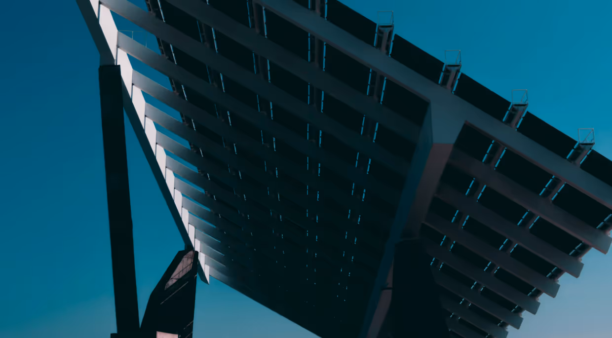 Low-angle view of a modern architectural structure featuring a geometric pergola or canopy with parallel wooden or metal slats creating a grid pattern of shadows, photographed against a clear teal-blue sky.