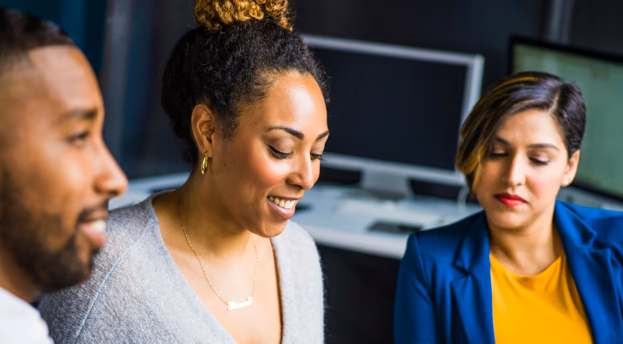 Alt text: "Three business professionals collaborating in a modern office environment with computer monitors in the background. A smiling woman with curly hair in a bun wearing a gray cardigan is in the center, flanked by a man on the left and a woman in a blue blazer and yellow top on the right.