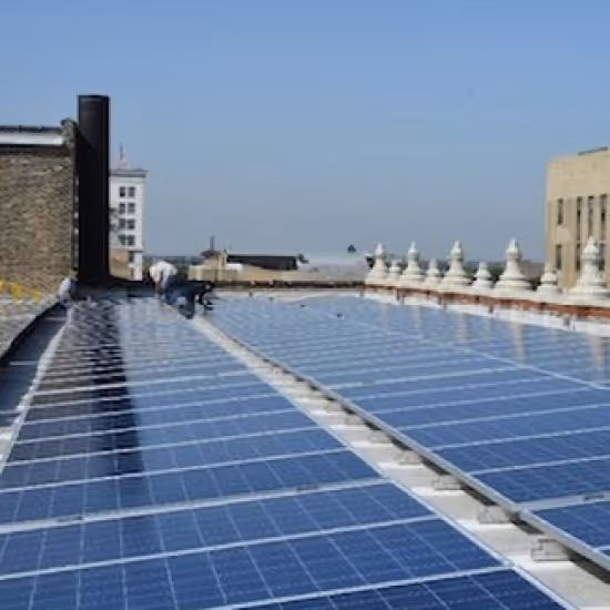 Rooftop solar panel installation on an urban building with blue photovoltaic panels arranged in rows across the flat roof, with city buildings and a clear blue sky visible in the background.