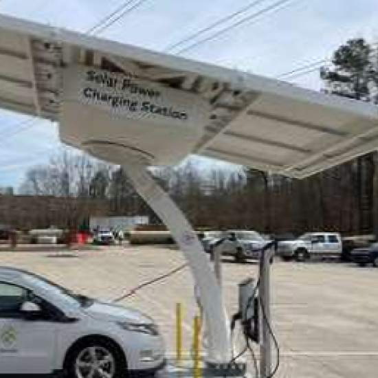 Solar-powered electric vehicle charging station with a white canopy featuring solar panels on top, positioned in a parking lot with a white car plugged in for charging and other vehicles visible in the background under an overcast sky.