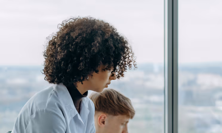 Woman with curly hair wearing a white button-up shirt looking down at a child with short hair, both positioned by a large window with a blurred cityscape visible in the background.