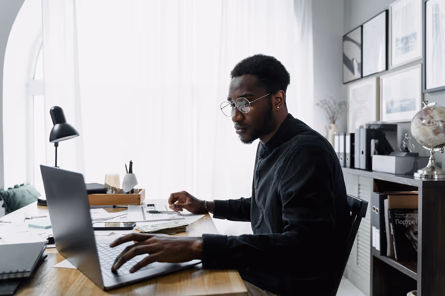 Man wearing glasses and a dark sweater working at a laptop computer at a wooden desk in a bright, modern home office with white shelving, framed artwork, a desk lamp, and a globe visible in the background.