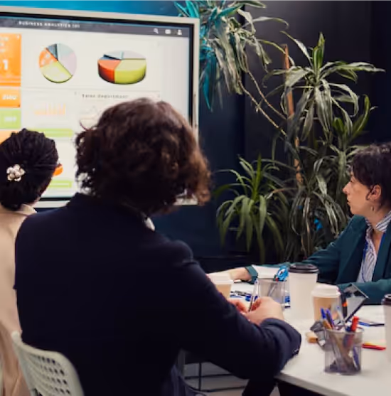Business meeting in a modern office with several people seated around a white table viewing a presentation on a wall-mounted screen displaying colorful charts and graphs, with a large plant visible in the background and office supplies on the table.