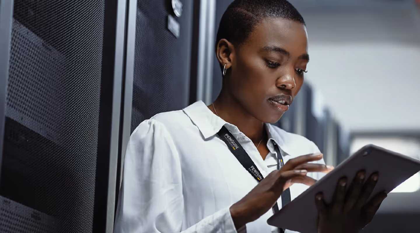Professional woman in a white collared shirt and black lanyard working on a tablet device in what appears to be a modern data center or server room environment with equipment racks visible in the background.
