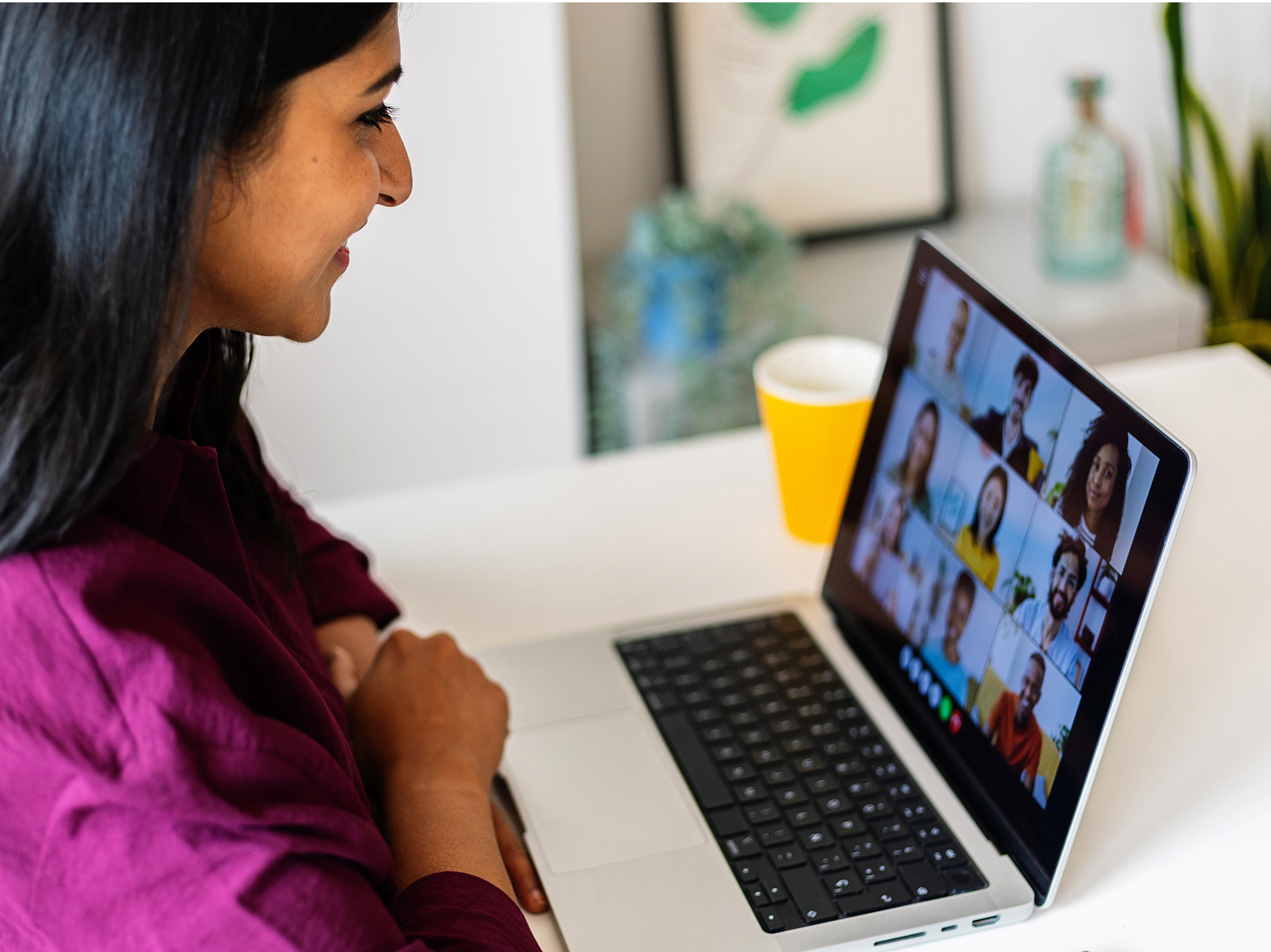 A woman with long dark hair wearing a burgundy/maroon top sits at a white desk, looking at a laptop screen displaying a video conference call with multiple participants in a grid layout. A yellow coffee mug sits on the desk next to the laptop.