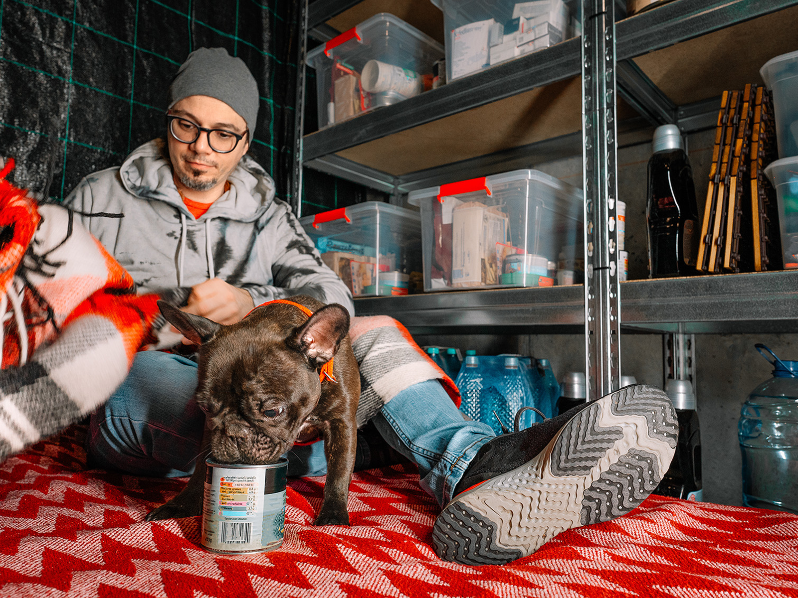 A man in glasses, gray hoodie, and beanie sits on a red patterned blanket in what appears to be a storage area or garage, gently petting a dark brown French bulldog. Behind them are metal shelving units containing clear storage containers and large water j