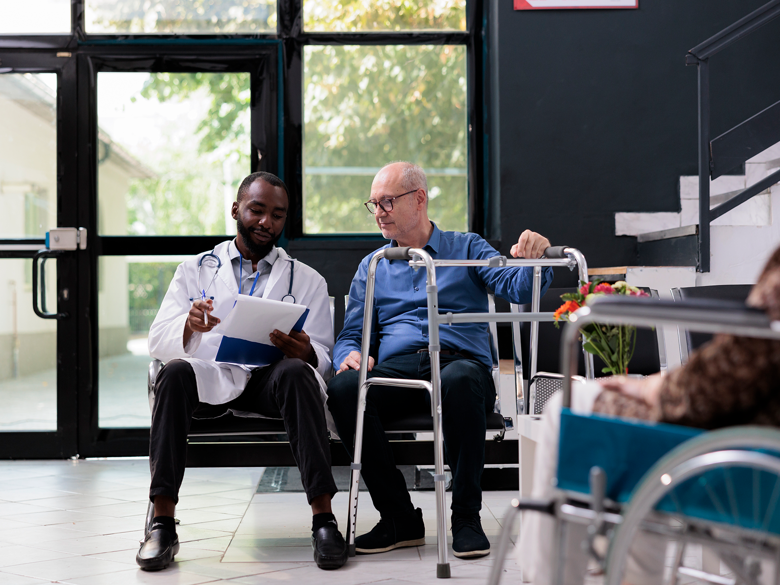 A doctor sits in an ER waiting room with a male patient using a walker.