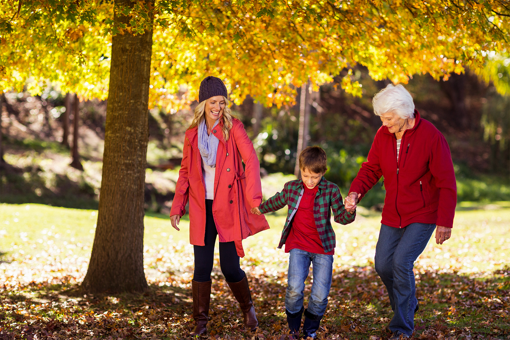 A mom, son, and grandmother are walking together in a park with fall leaves