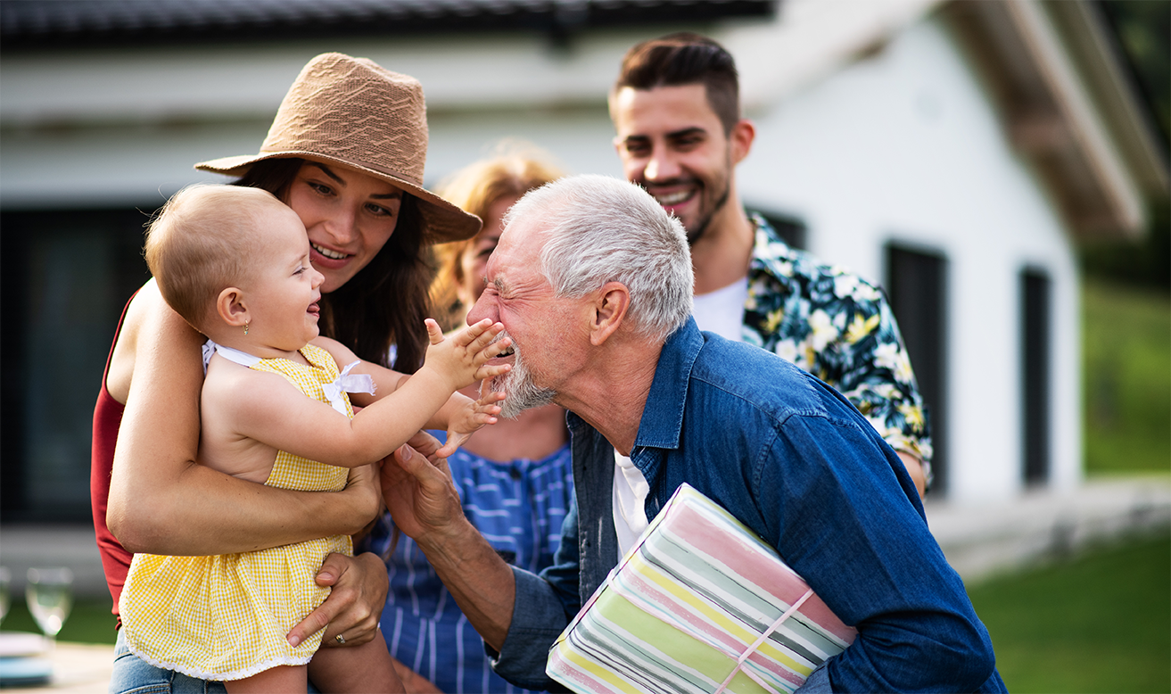 A multigenerational family is outside, where a young toddler is playfully reaching out for the face of a grandfather