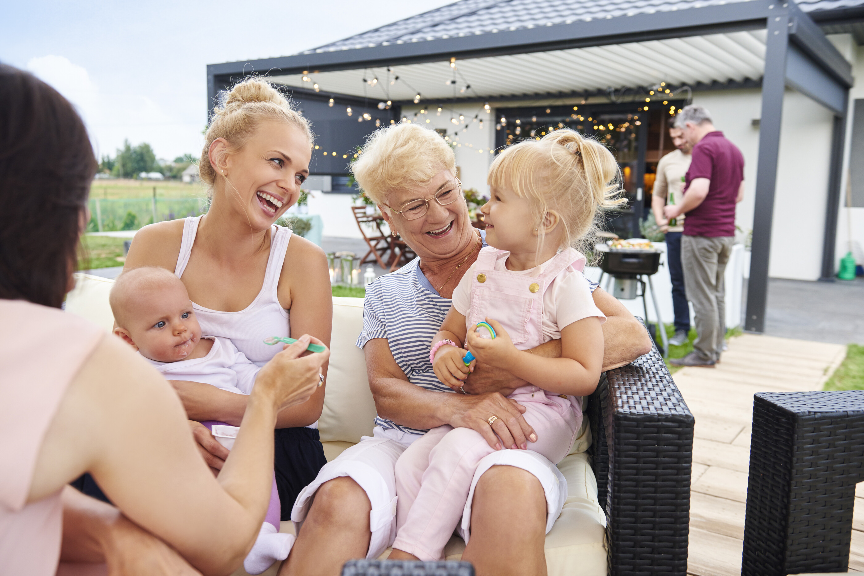 A multigenerational family is outside on a patio, with a mom, a young child, and a grandmother sitting on patio furniture 