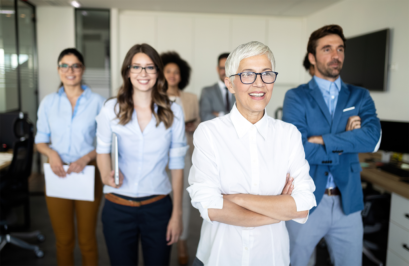 A group of coworkers of multiple generations stand together in an office