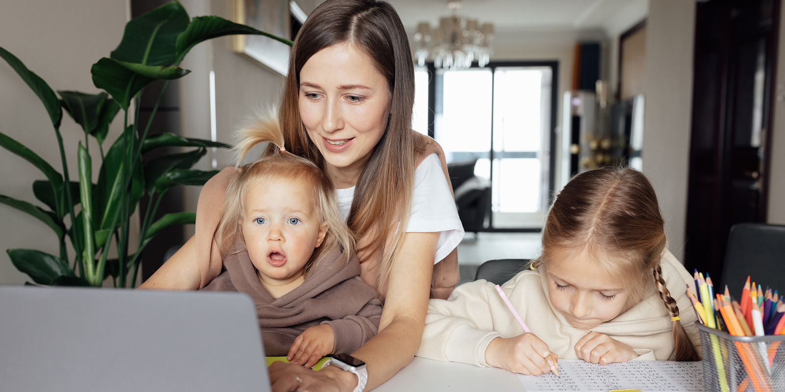A working mother types on a laptop at home while holding her toddler on her lap and her older daughter does schoolwork beside her at the kitchen table.