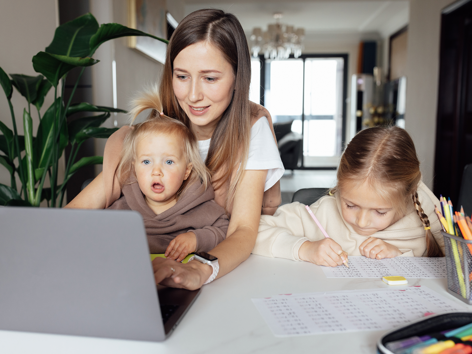 A working mother types on a laptop at home while holding her toddler on her lap and her older daughter does schoolwork beside her at the kitchen table.