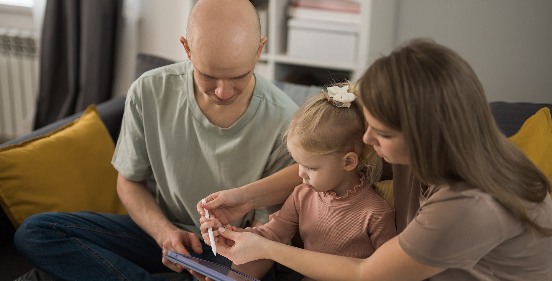 Two parents and their young child sitting together on a couch and looking at a tablet.
