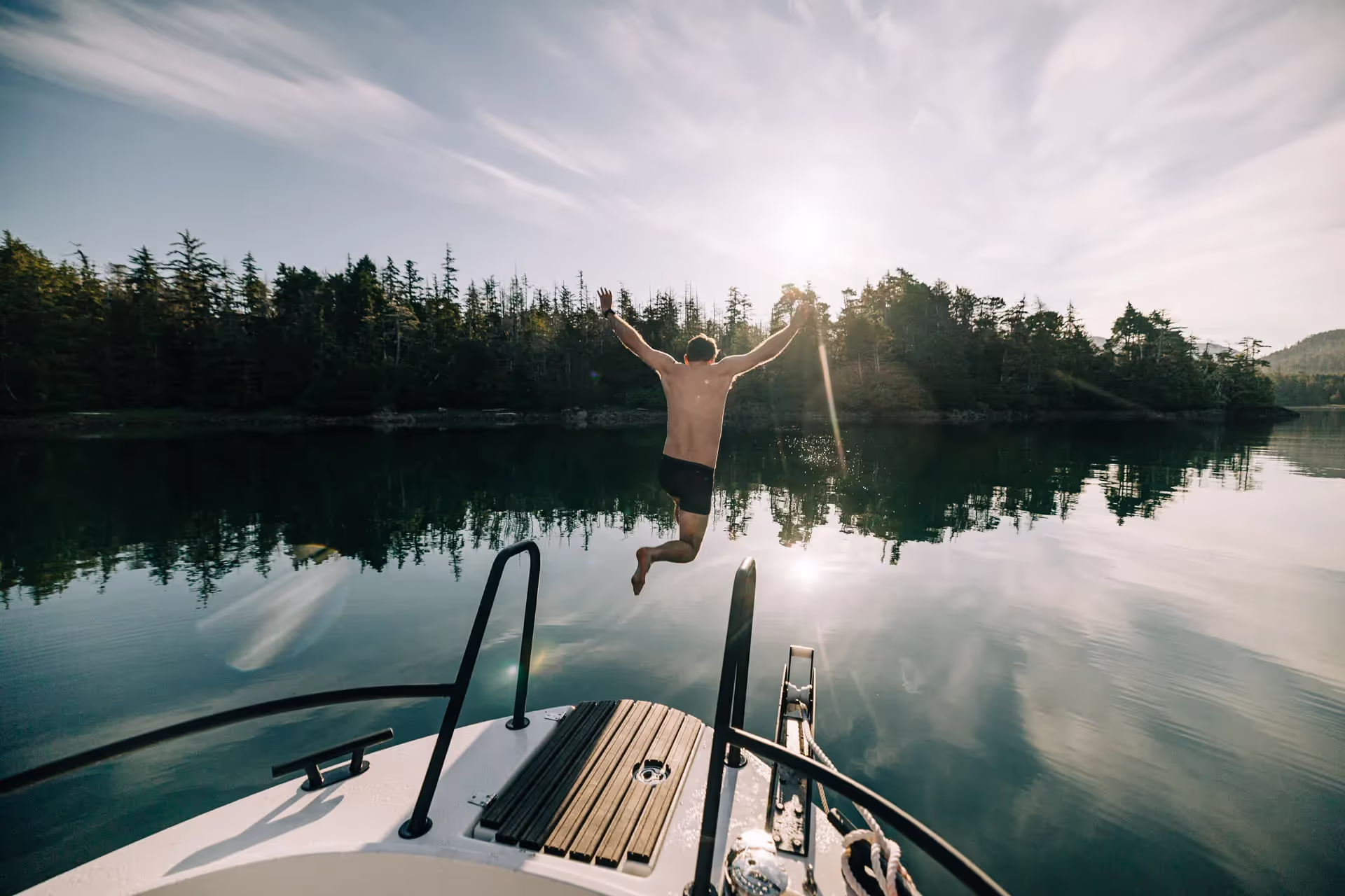 Man jumping off a boat into a calm bay with forested shore and sun low in the sky.