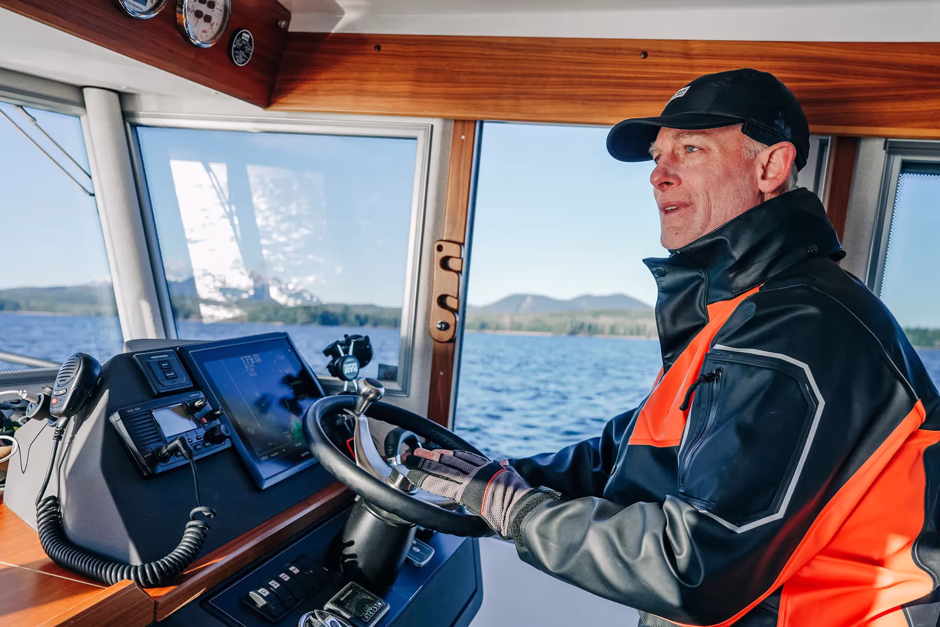 Man wearing a black cap and orange waterproof jacket steering a boat with ocean and mountains visible through the windows.